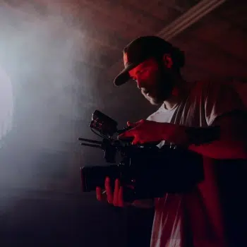 Persona con gorra sosteniendo una cámara de cine en un espacio oscuro con un foco de luz y humo