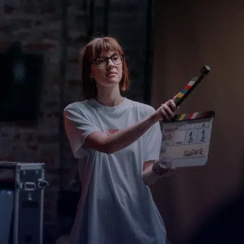 Mujer joven con gafas sostiene una claqueta frente a cámara en un set de rodaje, iluminada con luz tenue