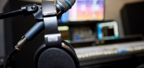 Micrófono y auriculares en primer plano y de fondo una mesa de sonido y la pantalla de un ordenador