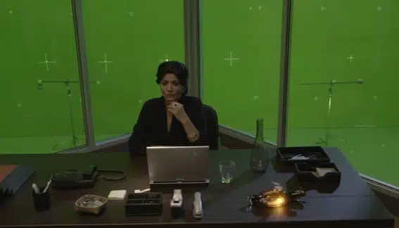 La imagen muestra la escena de un rodaje: una mujer está sentada en un escritorio y, a sus espaldas, hay un croma verde